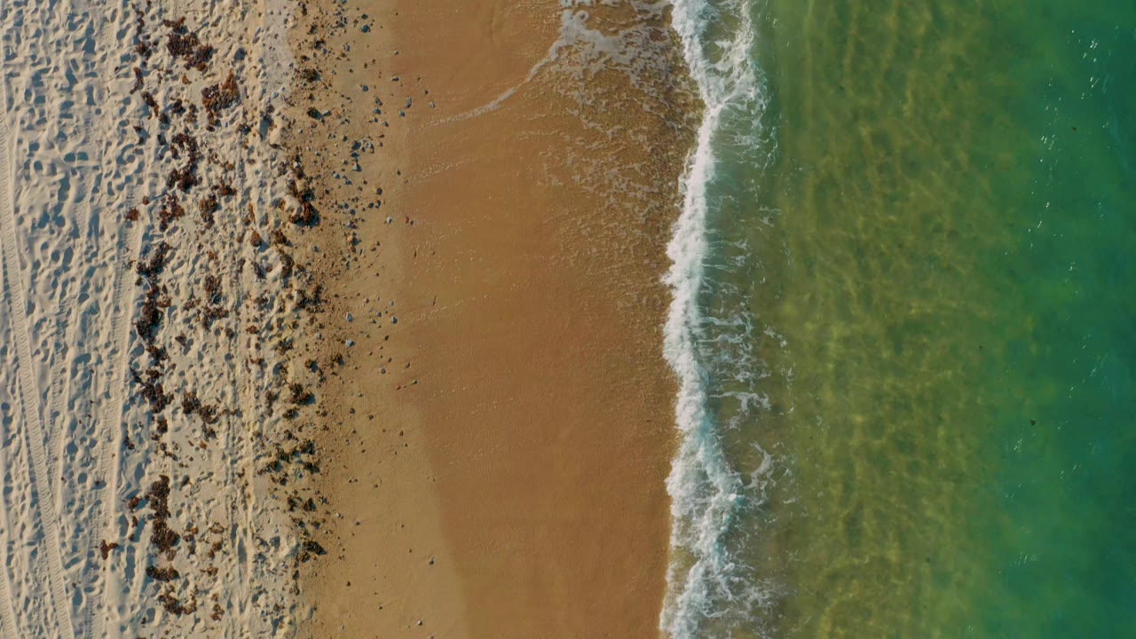 Top-down El Sargento La Ventana shoreline frame with beach tracks and scattered dark natural debris on the left, smooth tan sand in the middle, clear water on the right, and no visible people.