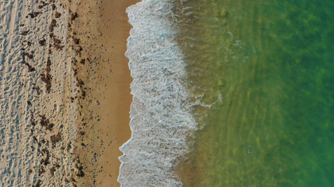 Overhead view in El Sargento La Ventana, Mexico, showing a diagonal band of white surf between tan sand and green water, with no visible people.