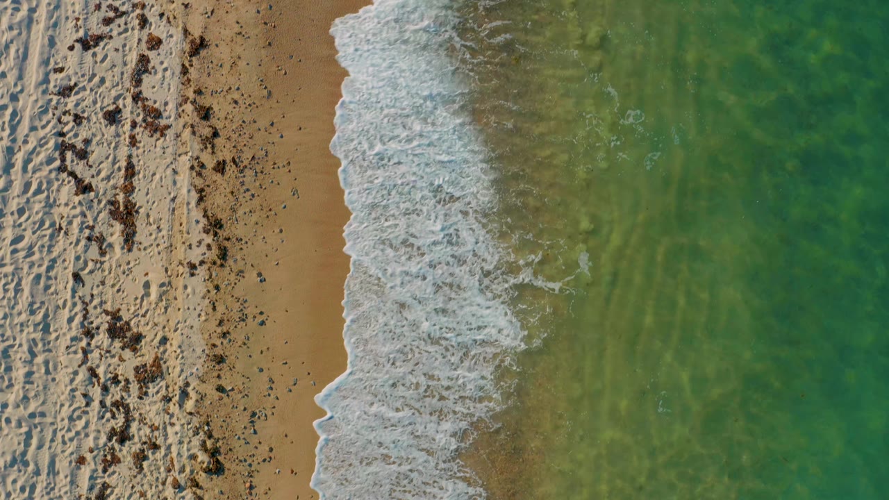 Aerial frame from El Sargento La Ventana, Mexico, with surf spread farther onto the wet sand and green water filling the right side, with no visible people.