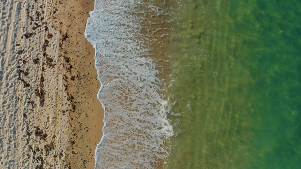 Top-down shoreline in El Sargento La Ventana, Mexico, where a curved white foam edge meets tan beach and green water, with no visible people.