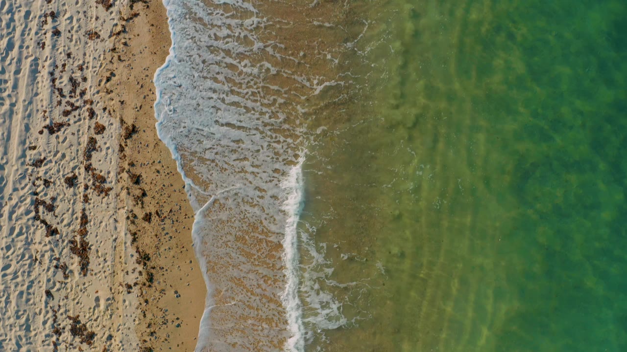 Overhead beach frame in El Sargento La Ventana, Mexico, showing a thinner foam line along the sand and translucent green water to the right, with no visible people.