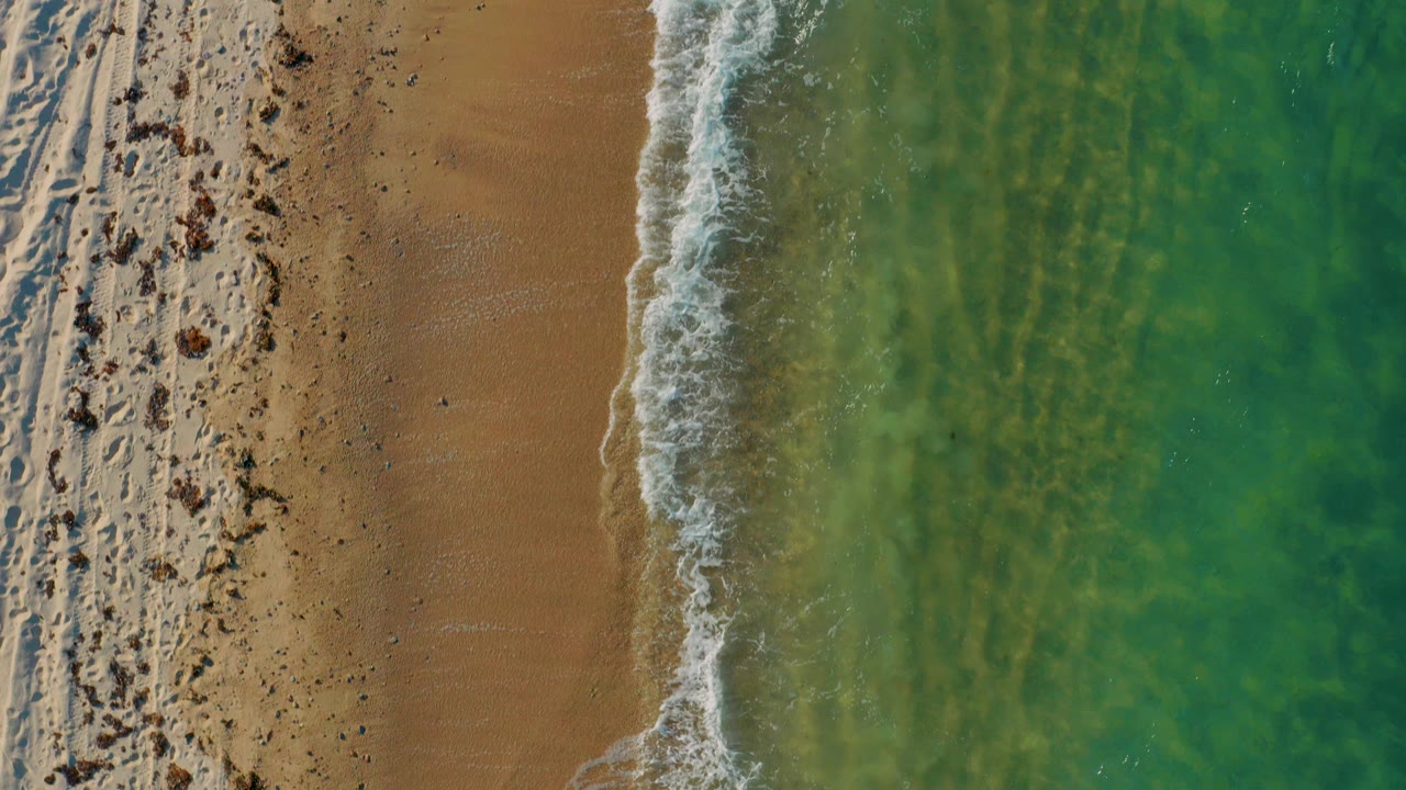 Top-down aerial frame of El Sargento La Ventana shoreline with tan sand at left, green-blue water at right, a thin surf line, scattered beach debris, and no visible people.