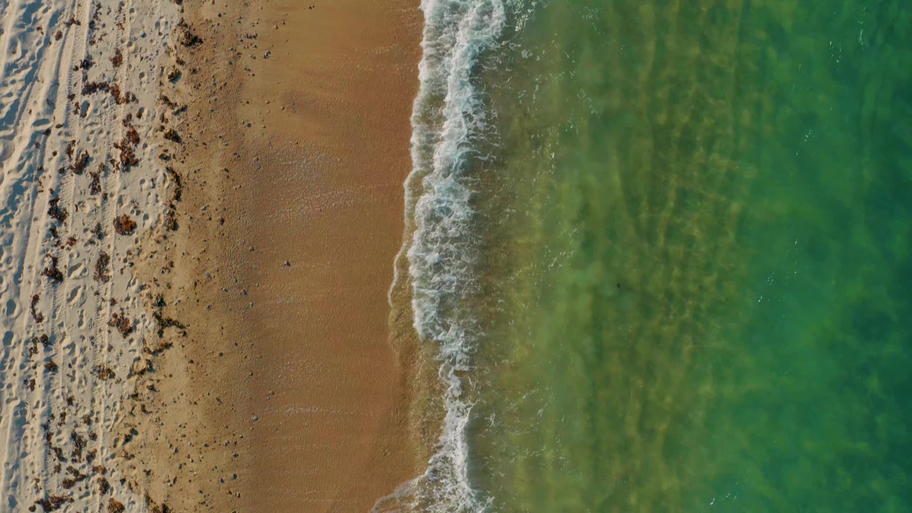 Aerial view over El Sargento La Ventana beach as the white foam line sits closer to the sand, with translucent green water and no visible people.