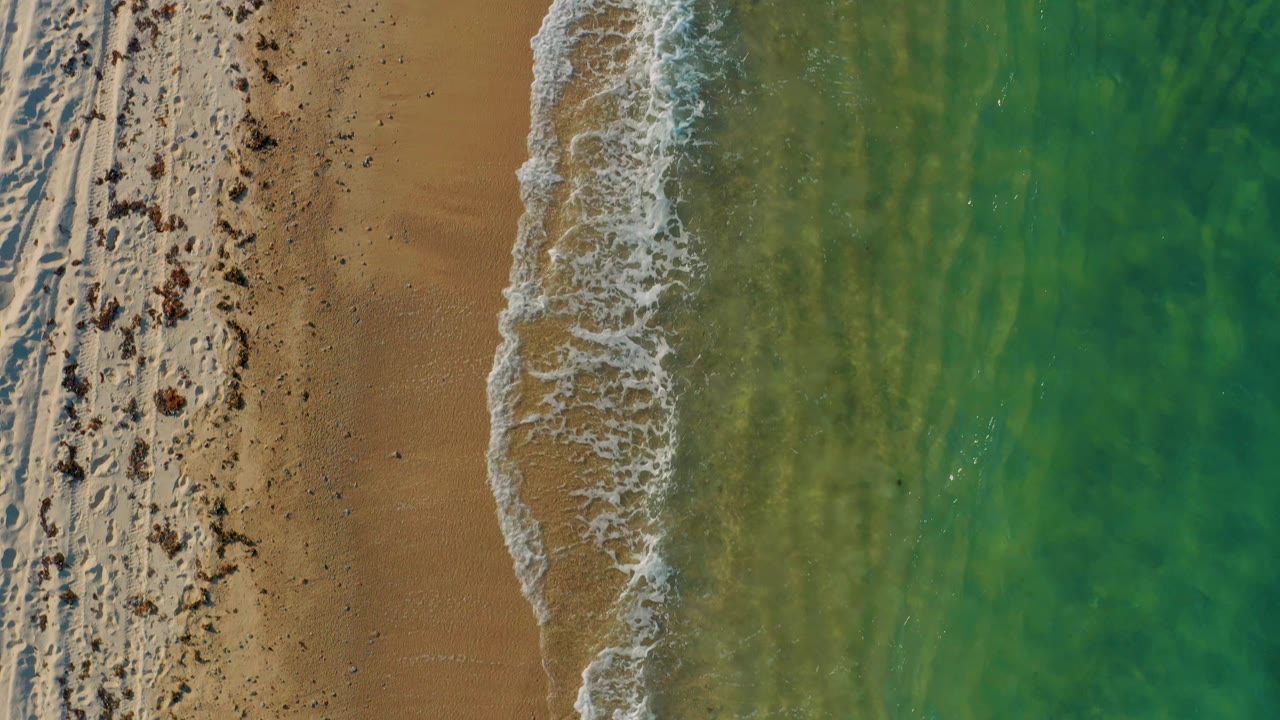 Top-down frame of the El Sargento La Ventana coast with a narrow tan beach band, white wash at the waterline, green ocean texture, and no visible people.