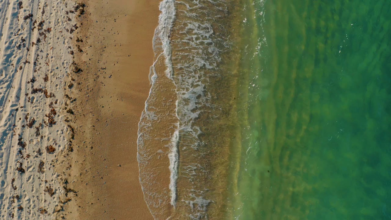 Aerial frame in El Sargento La Ventana showing foamy surf spreading over wet tan sand, with clear green water filling the right side and no visible people.