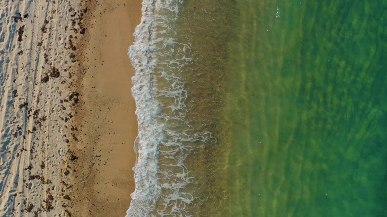 Overhead view of El Sargento La Ventana shoreline with tan sand on the left, green shallow water on the right, white surf between them, and no visible people.