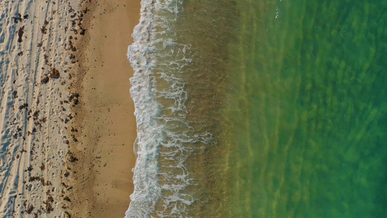 Aerial frame over El Sargento La Ventana showing a thicker white foam line curling along seaweed-marked sand beside green-blue water, with no visible people.
