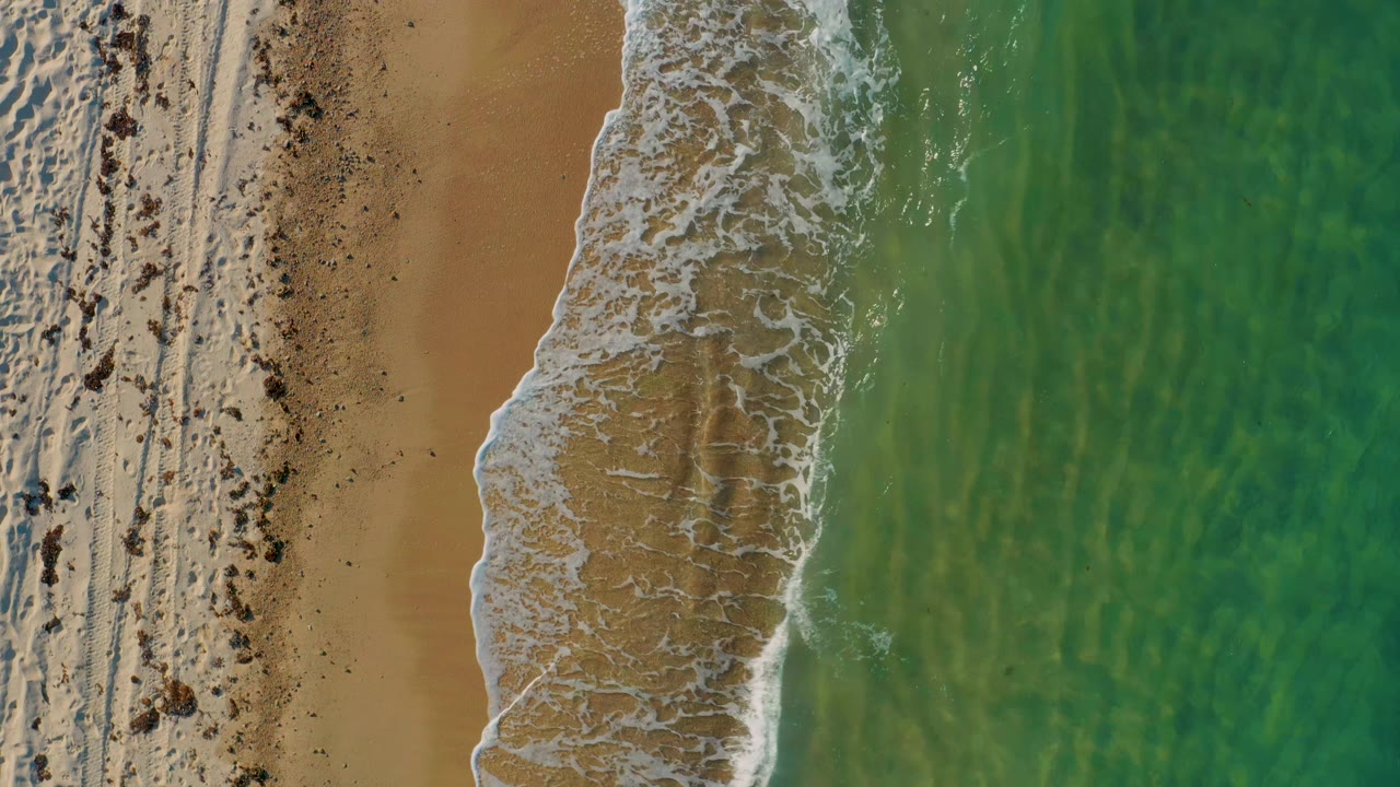 Aerial view in El Sargento La Ventana showing the foamy wave line pulled slightly lower along wet tan sand beside green water, with no visible people.