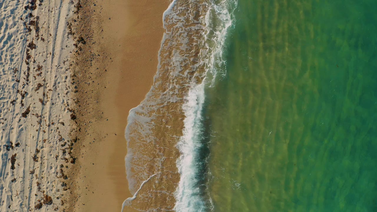 Top-down frame from El Sargento La Ventana with a stronger white breaker near the right side, tan beach to the left, translucent green water to the right, and no visible people.