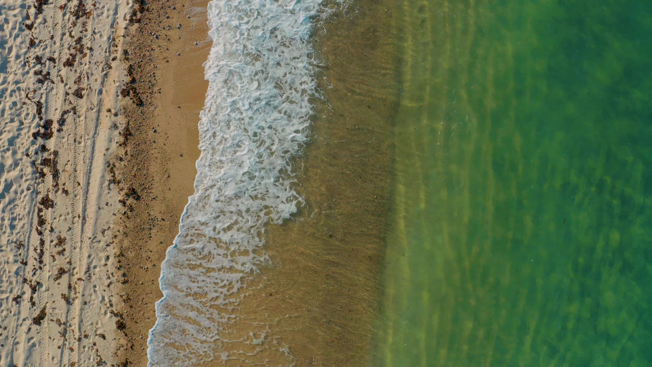 Overhead view of El Sargento La Ventana shoreline with dry textured sand on the left, a white foam band near center, turquoise water on the right, and no visible people.