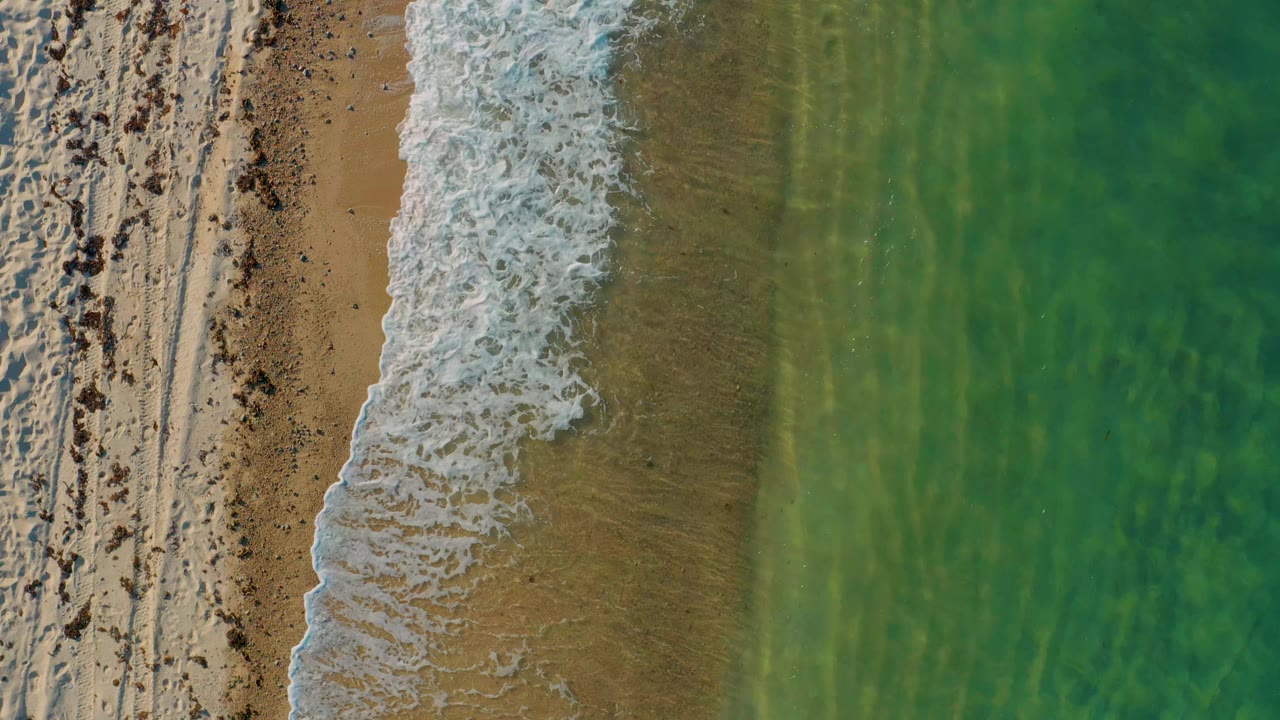 At 0:01, white surf has shifted along the vertical shoreline, with tan wet sand exposed beside clear green-blue water and no visible people.