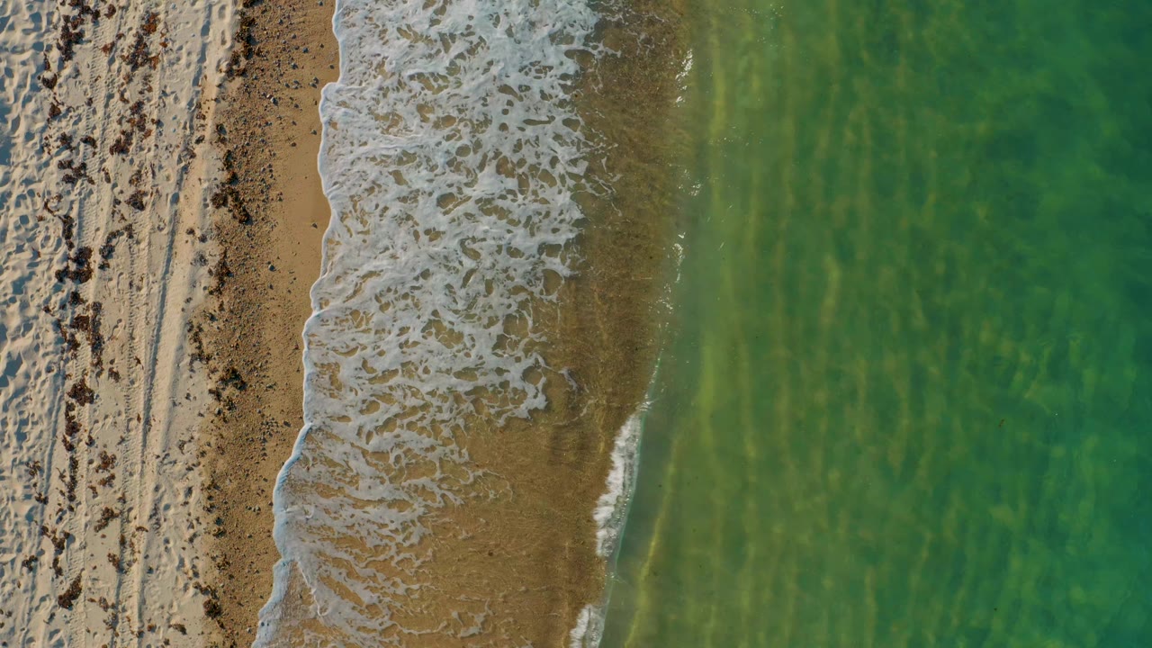 At 0:02, the overhead beach composition holds steady as a thicker patch of white foam crosses the center beside shallow turquoise water, with no visible people.