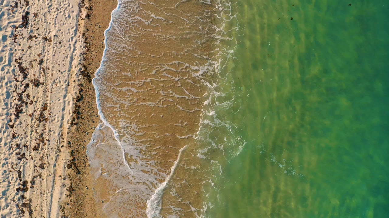 Aerial top-down view in El Sargento La Ventana, Mexico, with a curved wash of foam crossing tan wet sand and green water on the right, with no visible people.