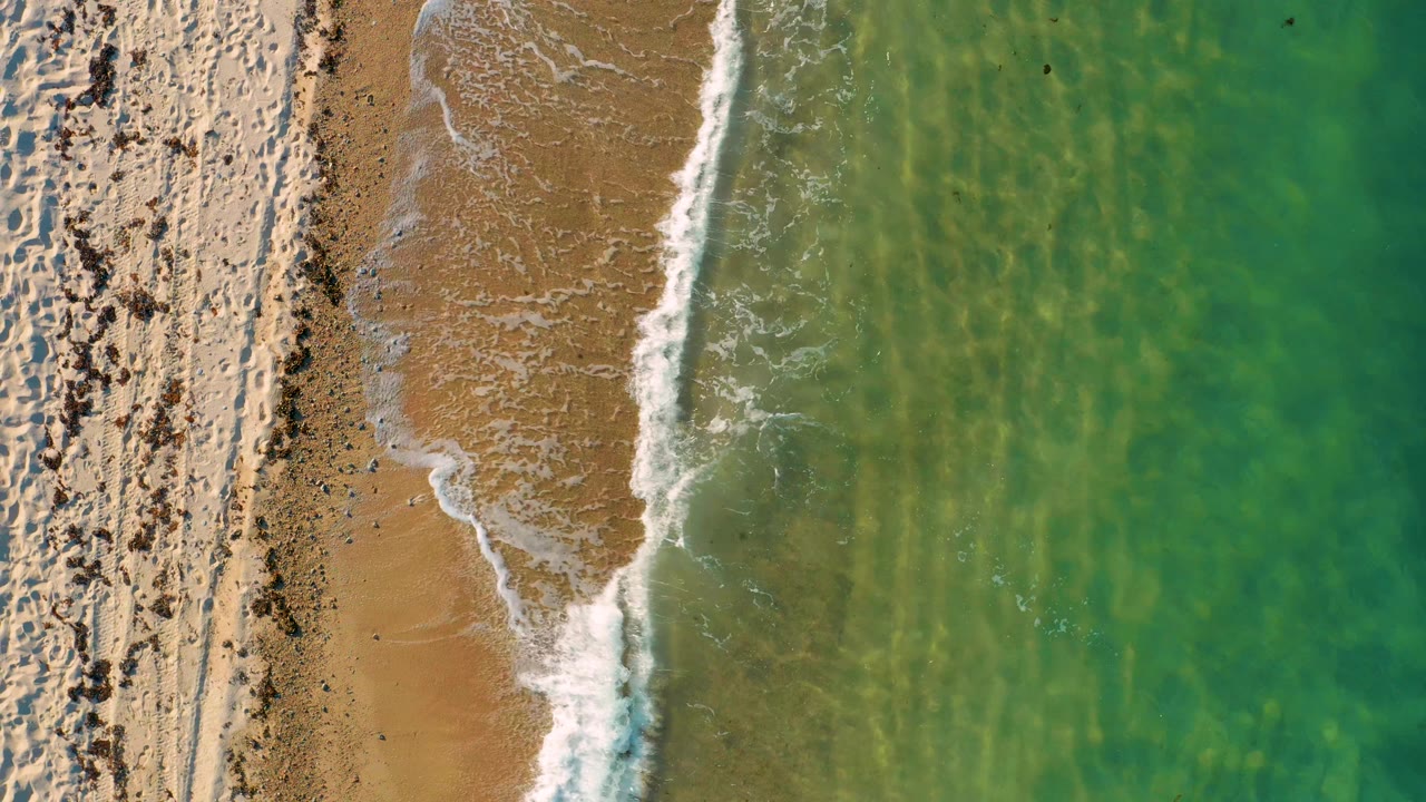 Straight-down aerial frame in El Sargento La Ventana, Mexico, showing a narrow sandy beach, a thin white wave line, and green water with faint underwater texture, with no visible people.