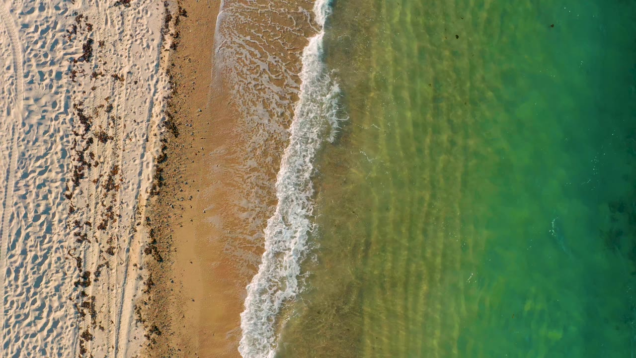 Aerial frame from El Sargento La Ventana, Mexico, with the surf line slightly farther along the beach, textured sand at left, translucent green water at right, and no visible people.
