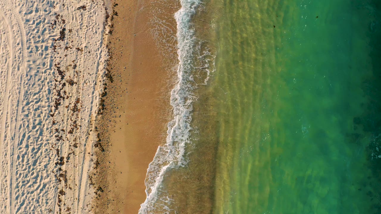Top-down frame from El Sargento La Ventana, Mexico, showing a broader curve of white surf against tan sand, green-blue water with darker areas, and no visible people.