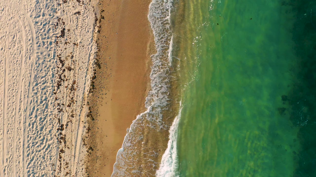 Overhead frame from El Sargento La Ventana, Mexico, where the beach and surf form a vertical strip, with rippled sand left, green water right, and no visible people.