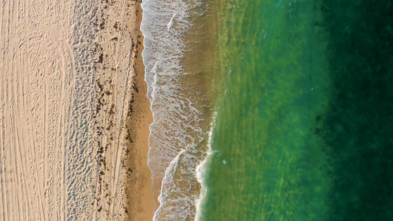 Overhead beach edge with pale sand, foam, and green water.