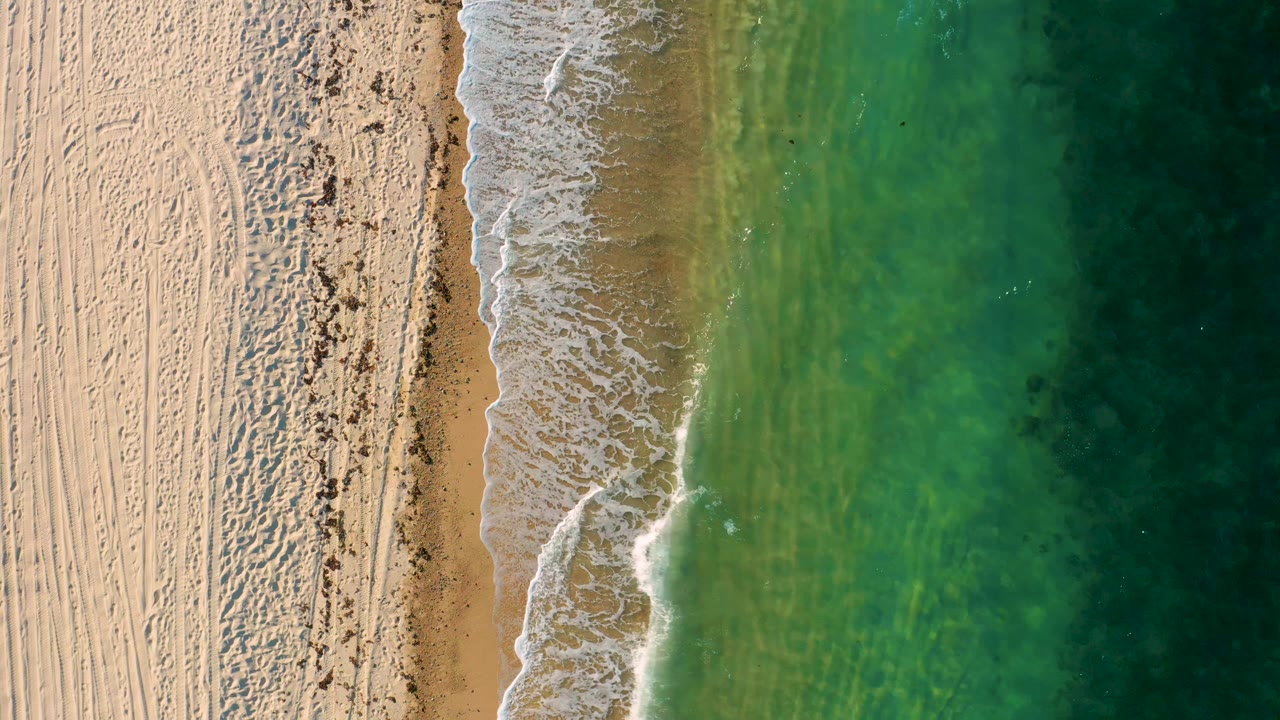 Overhead frame from El Sargento La Ventana showing a narrow surf band crossing the sand-water boundary, with translucent green water and no visible people.
