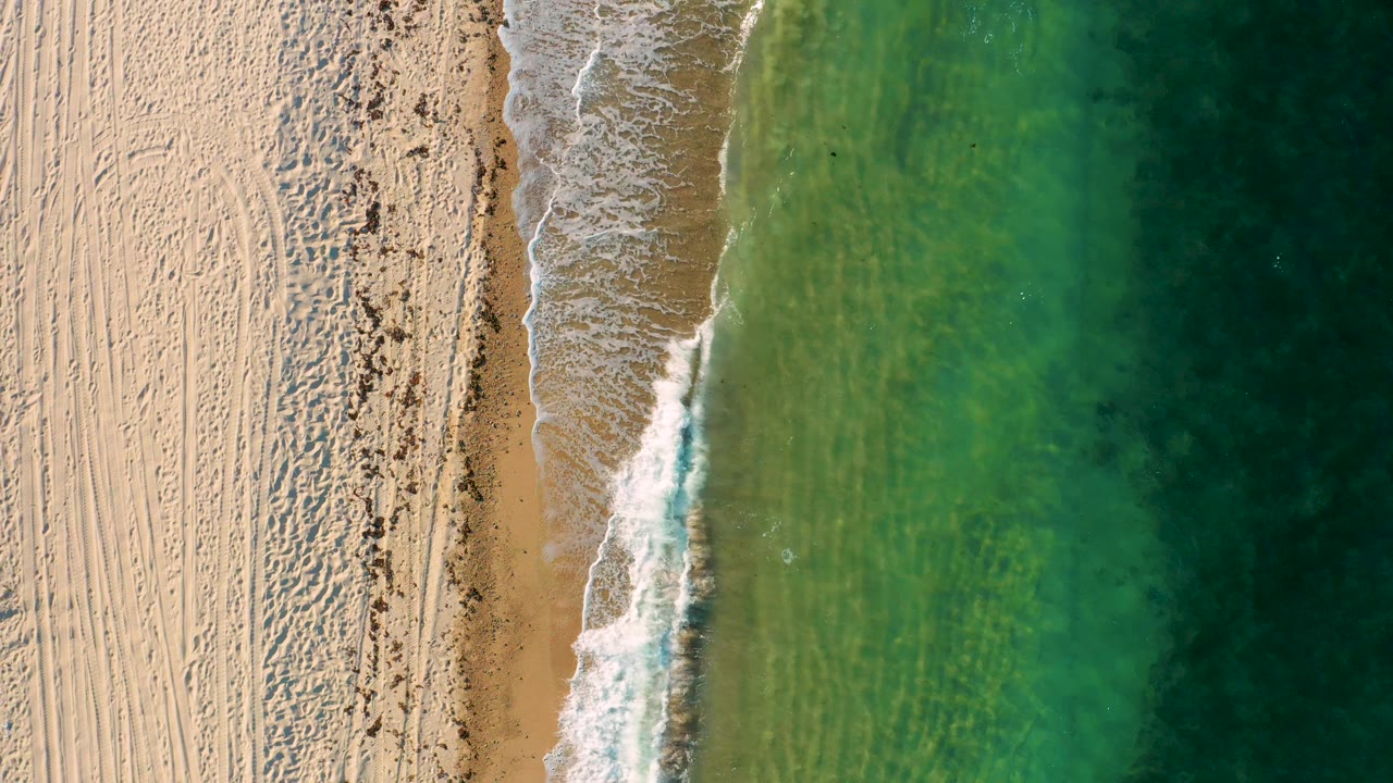 Top-down El Sargento La Ventana beach view with a thicker white wave band meeting brown wet sand, pale dry sand to the left, green water to the right, and no visible people.
