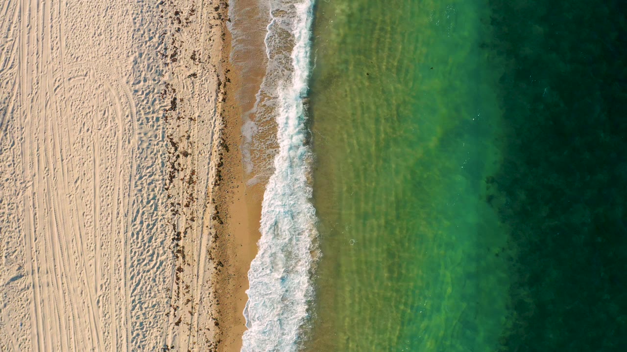Aerial frame over El Sargento La Ventana where a long white foam strip runs down the shoreline between textured sand and green water, with no visible people.