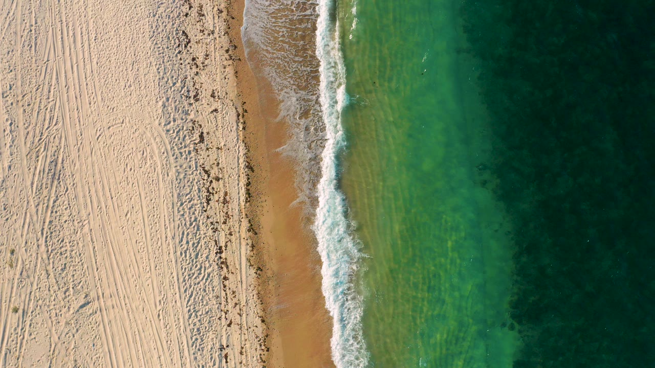 Top-down aerial frame of El Sargento La Ventana, Mexico, with pale sand on the left, a thin foamy wave line near center, green ocean on the right, and no visible people.