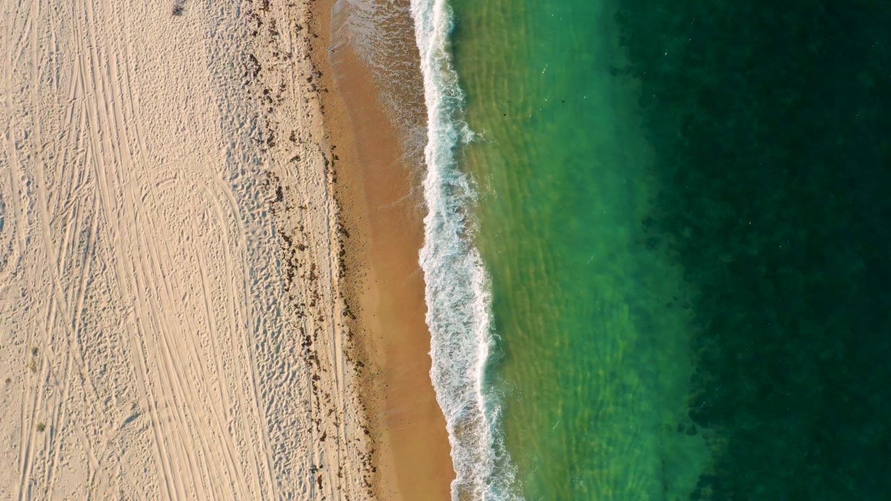 Top-down frame from El Sargento La Ventana, Mexico, with textured sand, a small dark object casting a shadow near the beach edge, foamy surf, green water, and no visible people.