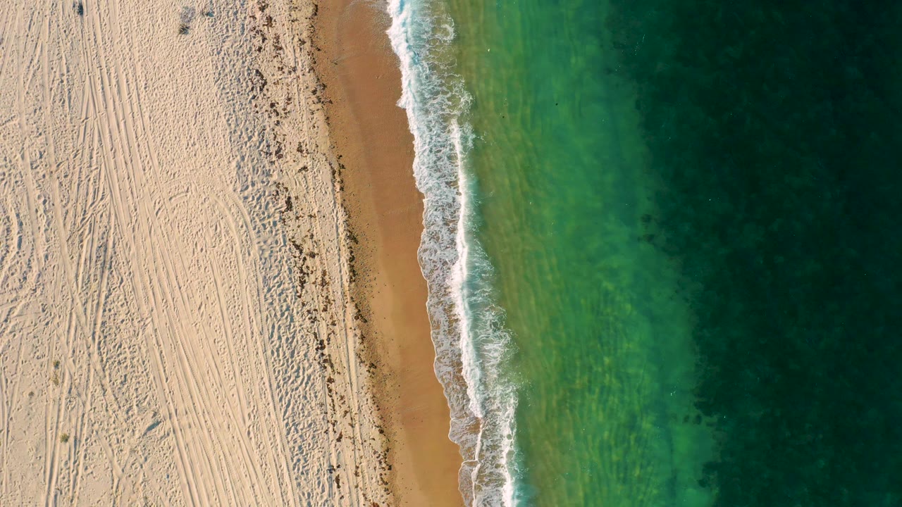 Aerial still over El Sargento La Ventana, Mexico, with empty pale sand on the left, a slim wet shoreline, white foam trailing along the beach, green ocean on the right, and no visible people.