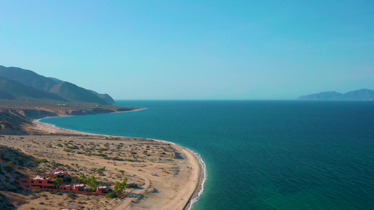 Opening aerial frame over El Sargento La Ventana, Mexico, showing a curved sandy beach, turquoise water, red village buildings at the lower left, mountains beyond, and no visible people.
