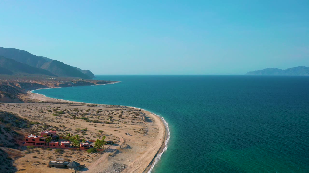 Aerial frame over El Sargento La Ventana, Mexico, showing the beach curve sweeping down the right side, red buildings and palms near the lower left, open sea across the right half, and no visible people.