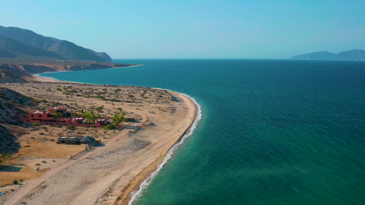 Aerial frame over El Sargento La Ventana, Mexico, with a sandy curved shore, turquoise water to the right, red buildings near the beach, mountains in the distance, and no visible people.