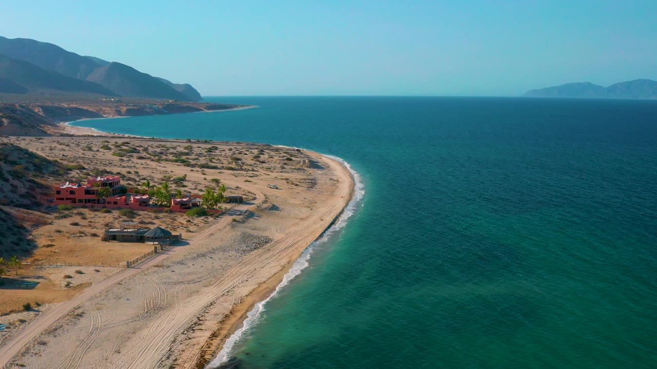 Aerial view over El Sargento La Ventana, Mexico, with more open sea on the right, a narrow sandy spit below, red structures inland from the beach, layered mountains on the left, and no visible people.