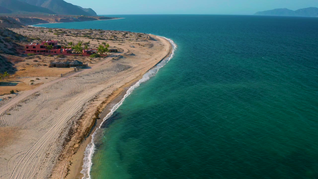 A high drone-like view in El Sargento La Ventana, Mexico, holds the beach curve while the surf traces the sand and red buildings sit near the hills, with no visible people.