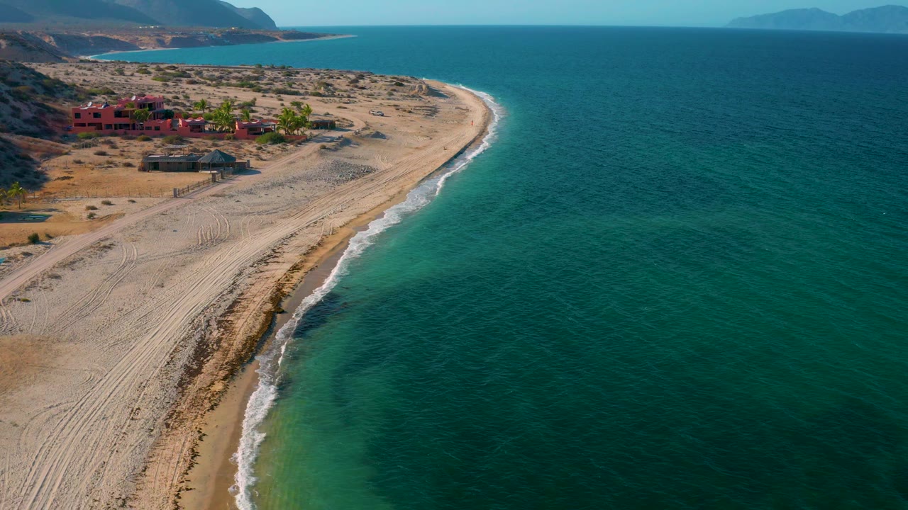 The frame shows El Sargento La Ventana shoreline from above, with more open water on the right, a pale beach edge on the left, distant mountains, red structures, and no visible people.
