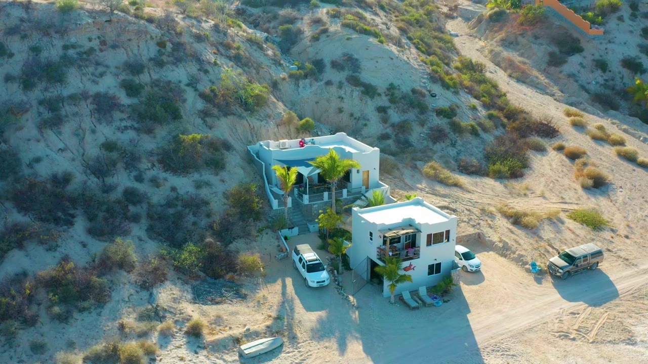 Bird's-eye frame of the white lodging buildings in El Sargento La Ventana, Mexico, with a white vehicle near the entrance, a second vehicle to the side, palms, desert brush, and no visible people.