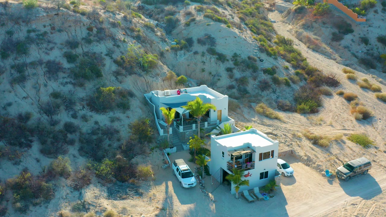 High-angle aerial frame over white modern homes, palm trees, parked vehicles, and sandy scrub hillside in El Sargento La Ventana, Mexico, with no visible people.