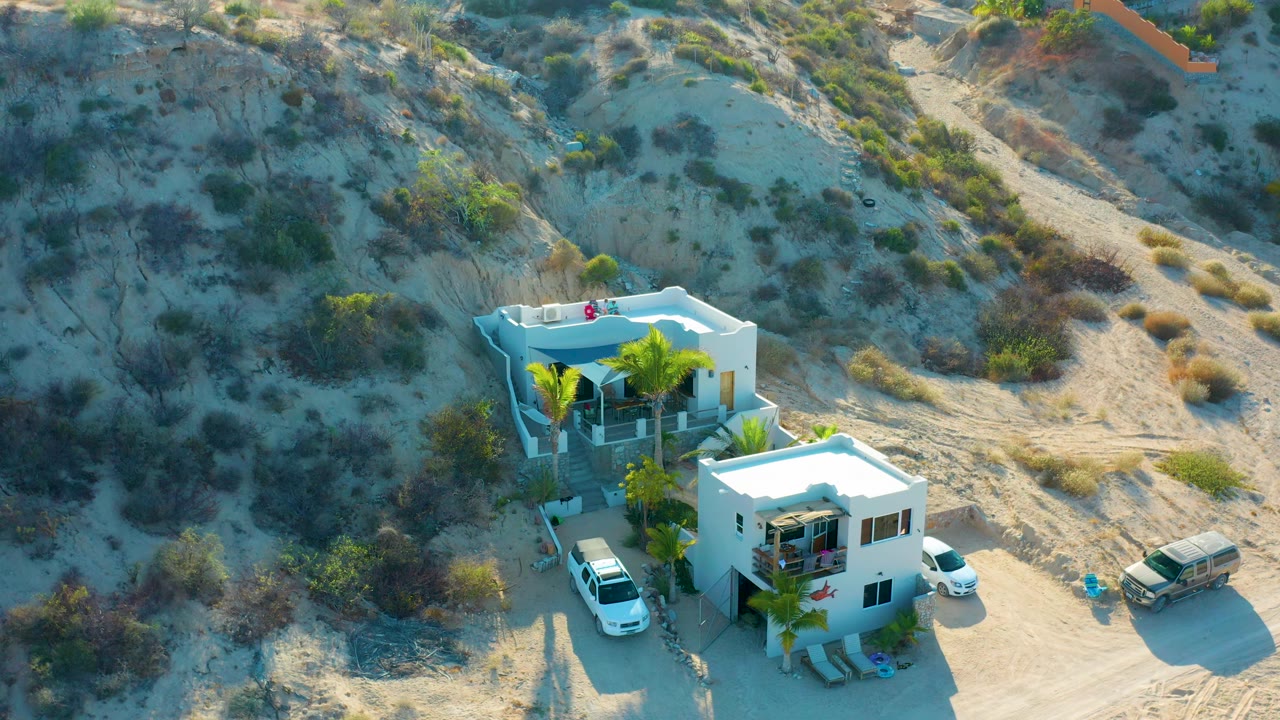Aerial frame looking down at modern white houses with palms and vehicles below pale sand ridges and low brush in El Sargento La Ventana, Mexico, with no visible people.
