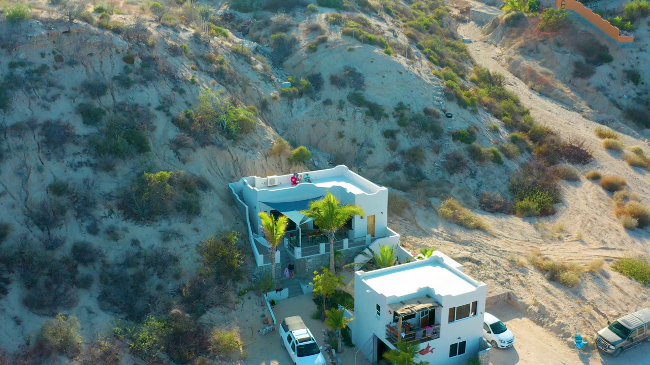 Opening overhead frame in El Sargento La Ventana, Mexico, showing two white modern houses, palm trees, parked cars, sandy hillside scrub, and no visible people.