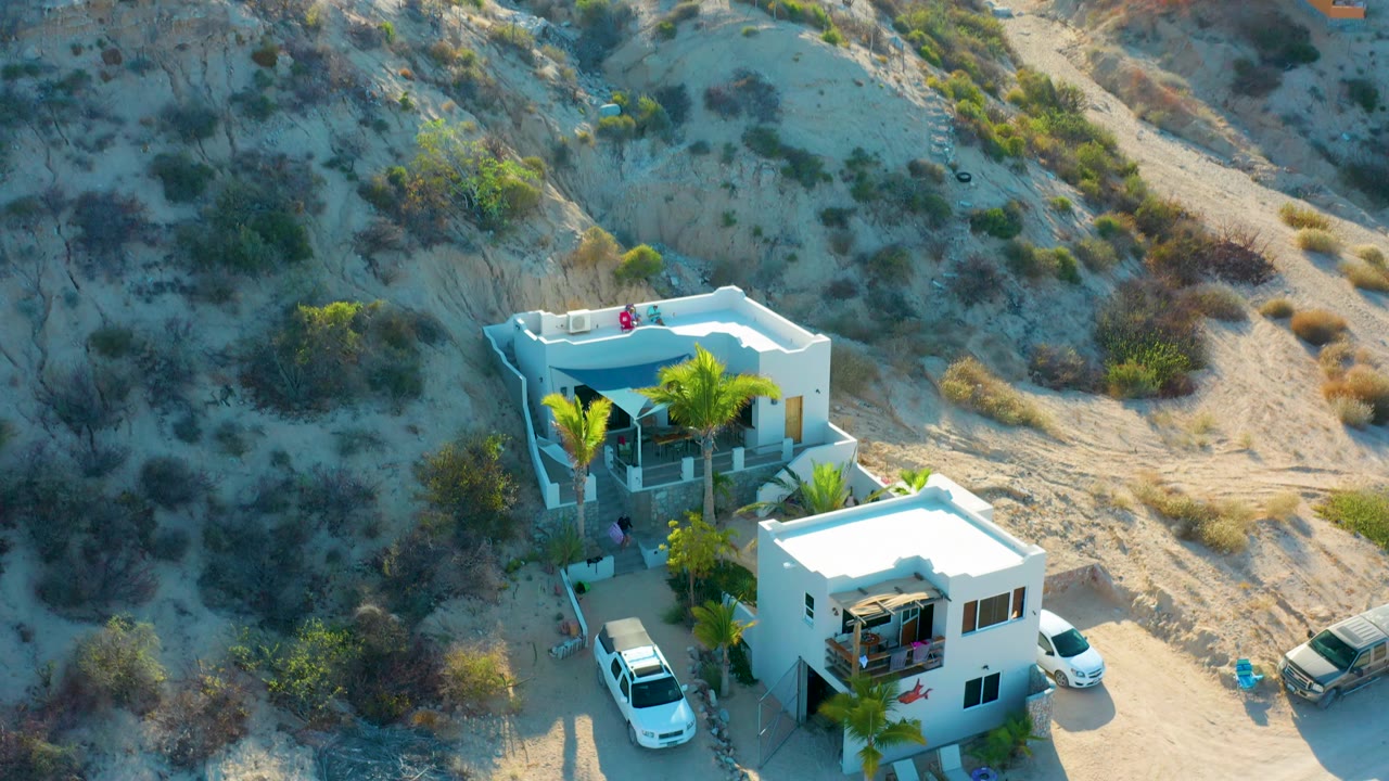 High aerial frame in El Sargento La Ventana, Mexico, showing flat-roof white houses, a white vehicle beside one home, a tan vehicle on the road, dry brush, and no visible people.