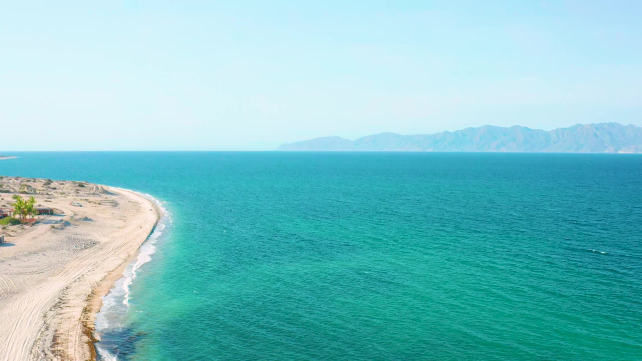 Aerial view over El Sargento La Ventana, Mexico, with a curved sandy shore on the left, turquoise sea across the frame, distant land on the horizon, and no visible people.