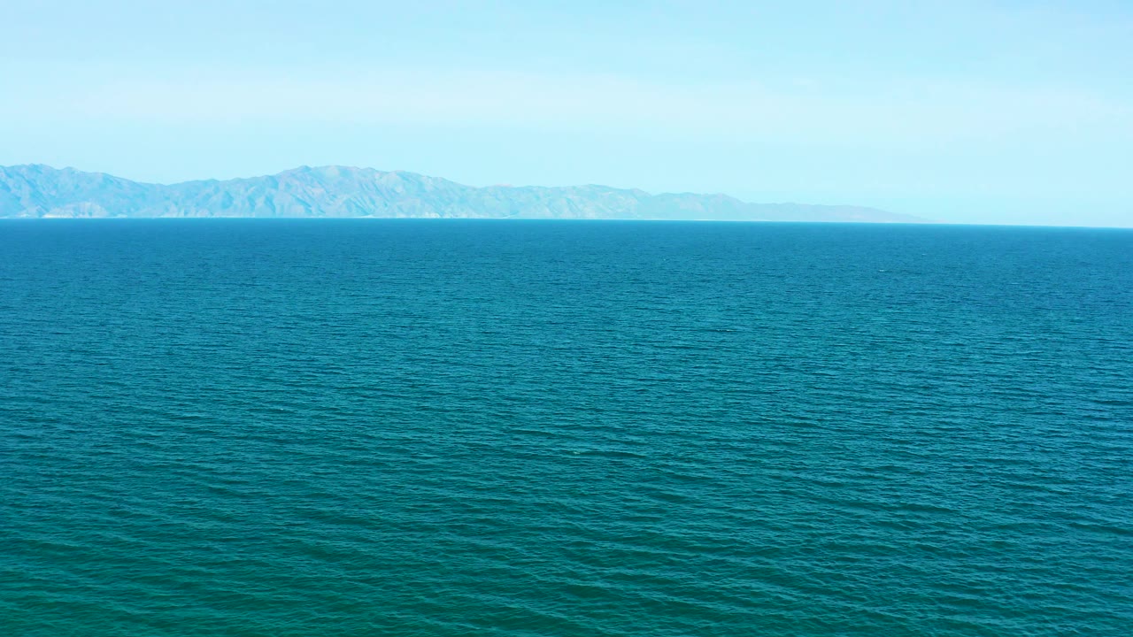 A similar wide frame holds more open blue-green water in the foreground with the mountain coastline centered along the horizon, no visible people.
