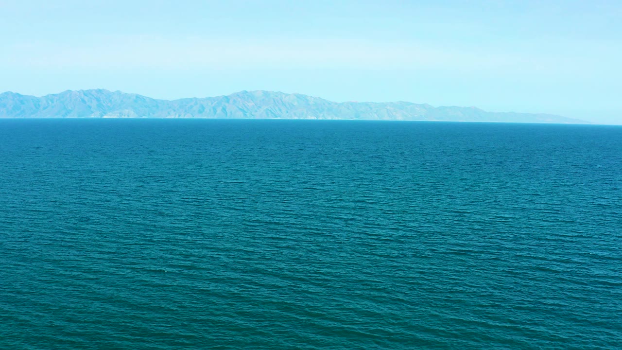 Opening frame of the El Sargento La Ventana, Mexico clip showing textured teal ocean, a straight horizon, distant mountainous land, pale sky, and no visible people.