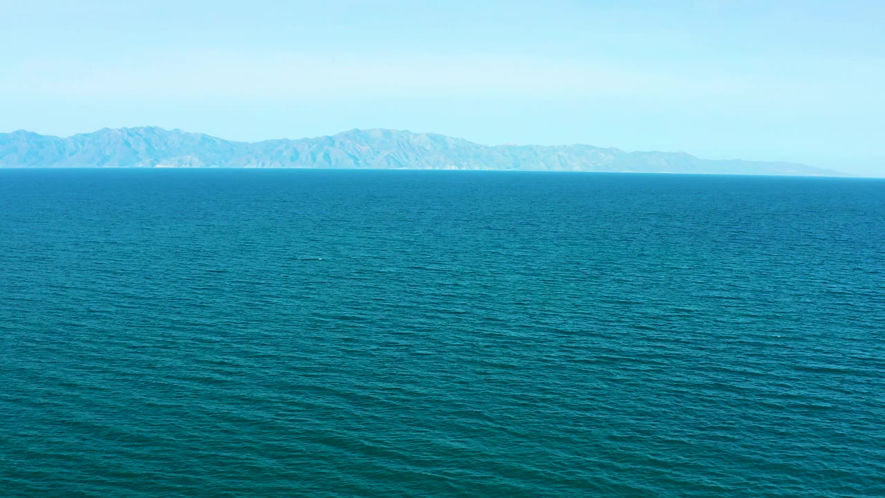At two seconds, the frame holds on a broad teal water foreground with a hazy mountainous island or coastline in the background, bright sky, and no visible people.