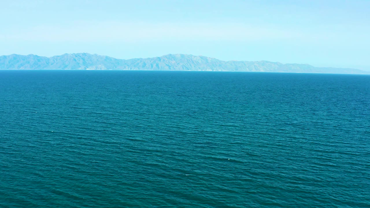 Aerial ocean frame from El Sargento La Ventana, Mexico, showing textured blue water below and hazy mountains stretching across the far horizon with no visible people.