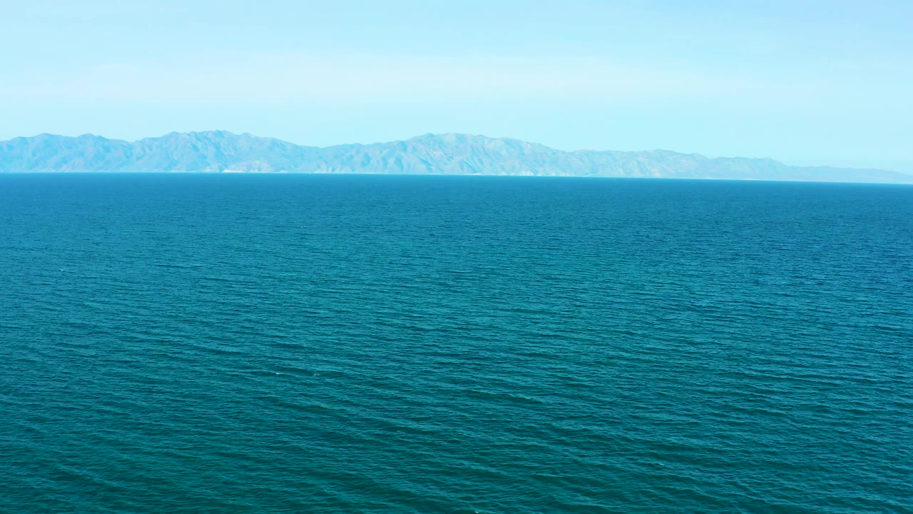 Wide aerial view near El Sargento La Ventana, Mexico, with open blue water in front and a low mountain shoreline running behind it, with no visible people.