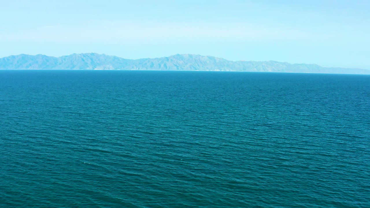 Aerial frame over blue water at El Sargento La Ventana, Mexico, with distant rocky coastline more defined along the horizon and no visible people.