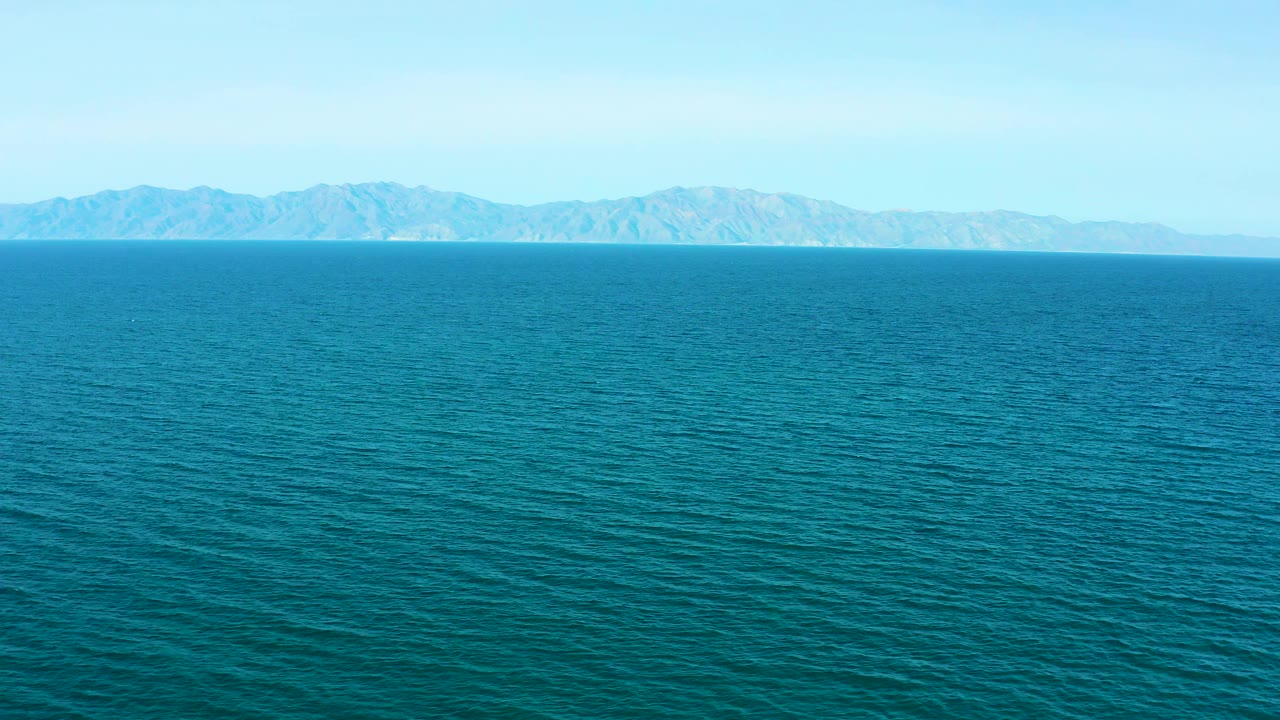 Aerial frame of textured blue-green sea leading to a long hazy mountain range across the horizon in El Sargento La Ventana, Mexico, with no visible people.