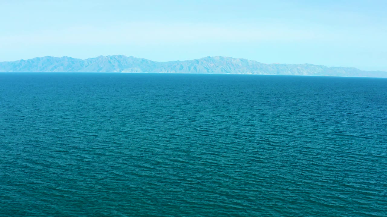 Wide coastal aerial over rippled blue ocean, with hazy rugged mountains stretching across the upper frame in El Sargento La Ventana, Mexico, and no visible people.