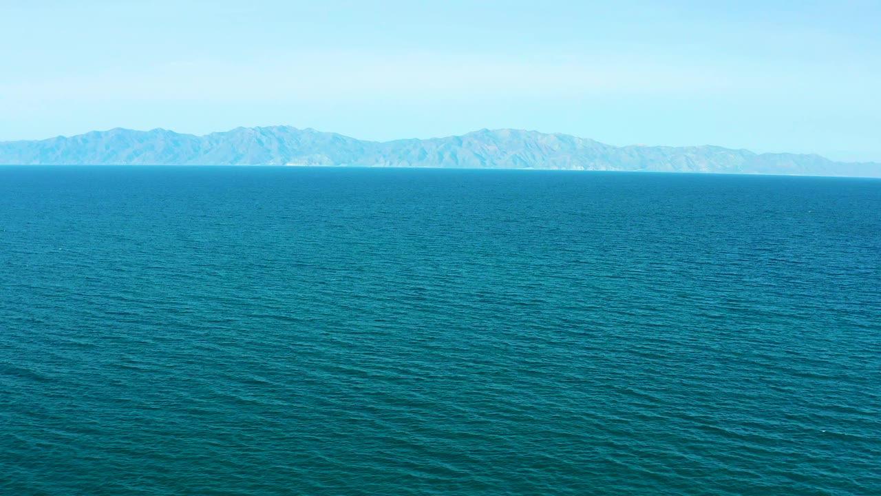 Bright aerial view across open turquoise sea toward low distant mountains and a clear sky in El Sargento La Ventana, Mexico, with no visible people.