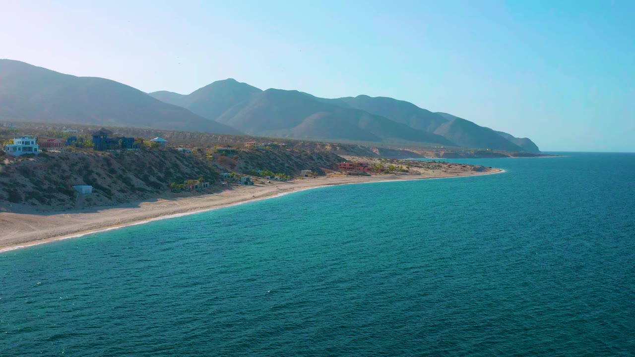 Opening aerial frame over El Sargento La Ventana, Mexico, with teal ocean, a narrow sandy beach, low coastal buildings, blue mountains, clear sky, and no visible people.